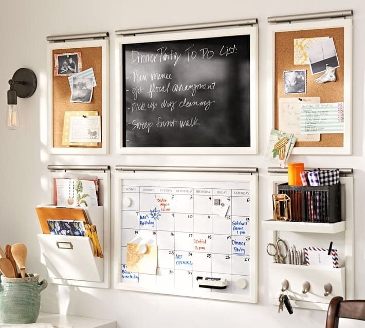 A modular white-framed wall command center featuring a chalkboard to-do list, a monthly whiteboard calendar, cork bulletin boards, hanging mail organizers, and a shelf with key hooks.