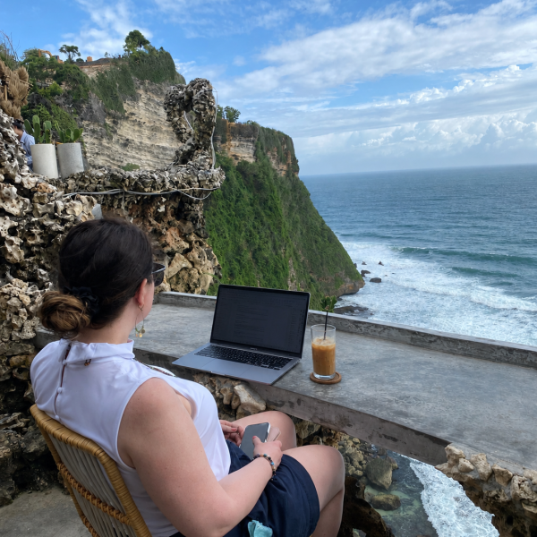 Marybeth Welty practicing work and travel organization for women entrepreneurs, sitting at an open-air cliffside desk in Bali with a laptop and iced coffee overlooking the ocean.
