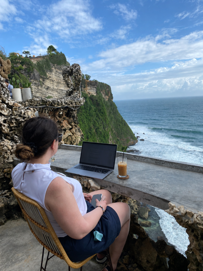 Marybeth Welty practicing work and travel organization for women entrepreneurs, sitting at an open-air cliffside desk in Bali with a laptop and iced coffee overlooking the ocean.
