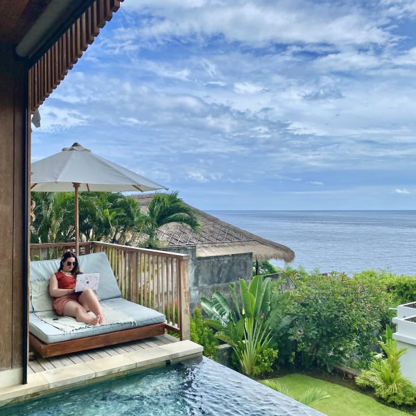 Woman entrepreneur working on laptop from a luxury Bali villa daybed beside a private infinity pool overlooking the ocean