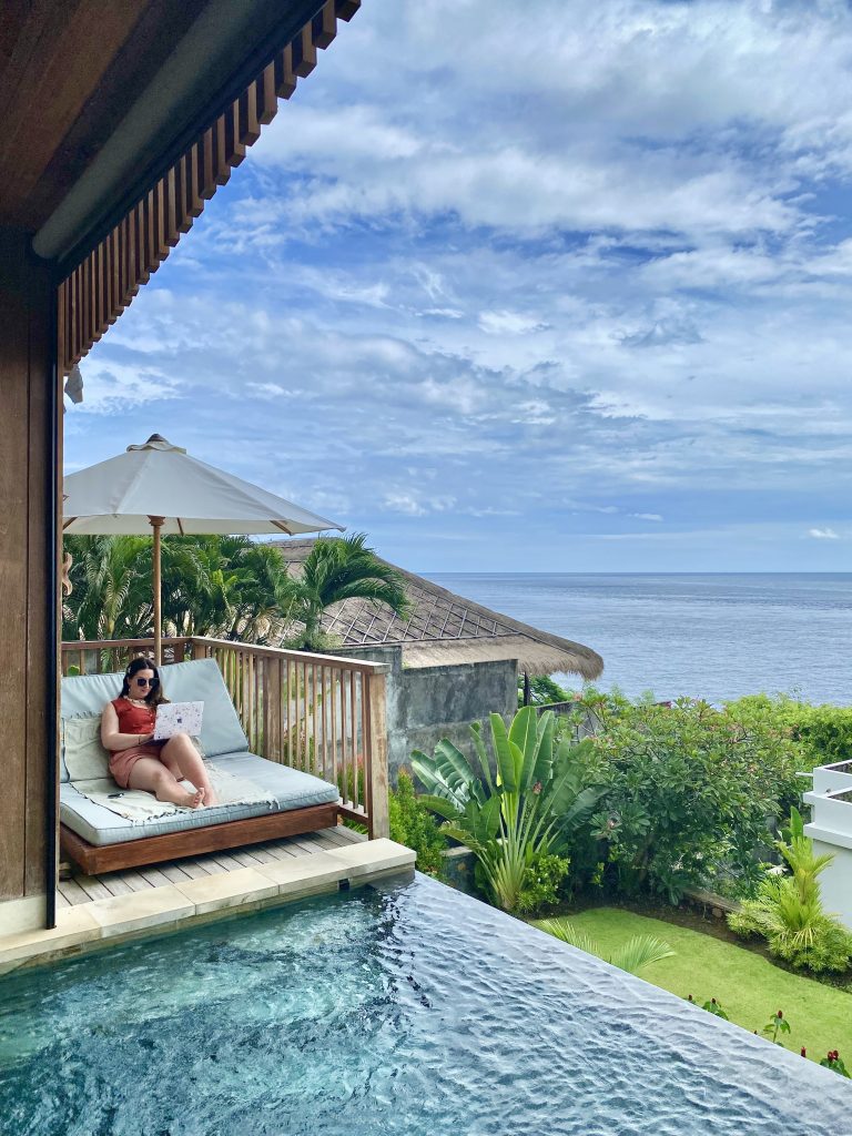 Woman entrepreneur working on laptop from a luxury Bali villa daybed beside a private infinity pool overlooking the ocean