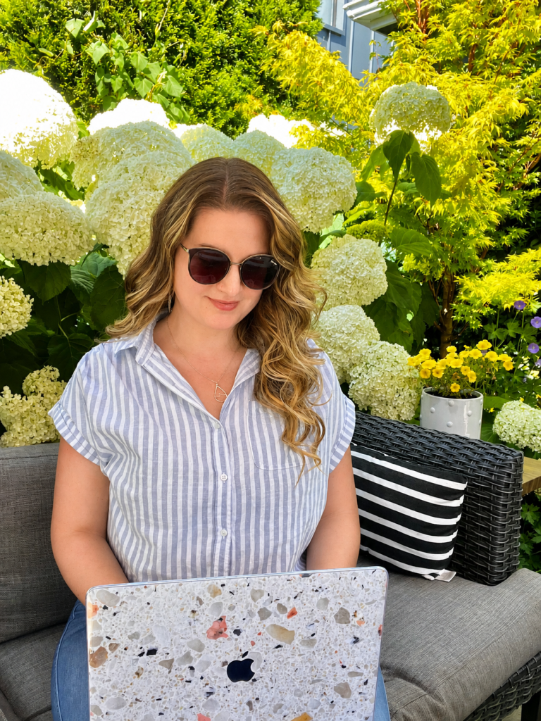 A woman wearing sunglasses and a striped shirt works on a laptop with a terrazzo-patterned skin, sitting on a grey wicker couch in a dense garden with large white hydrangeas, representing the freedom of a flexible, automated business.