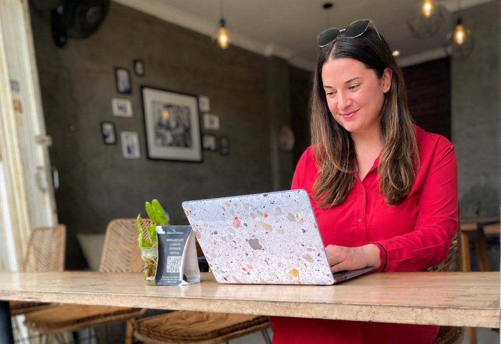 A woman entrepreneur working on her laptop at a wooden table in a lush, tropical cafe in Bali, surrounded by green plants and a peaceful atmosphere.