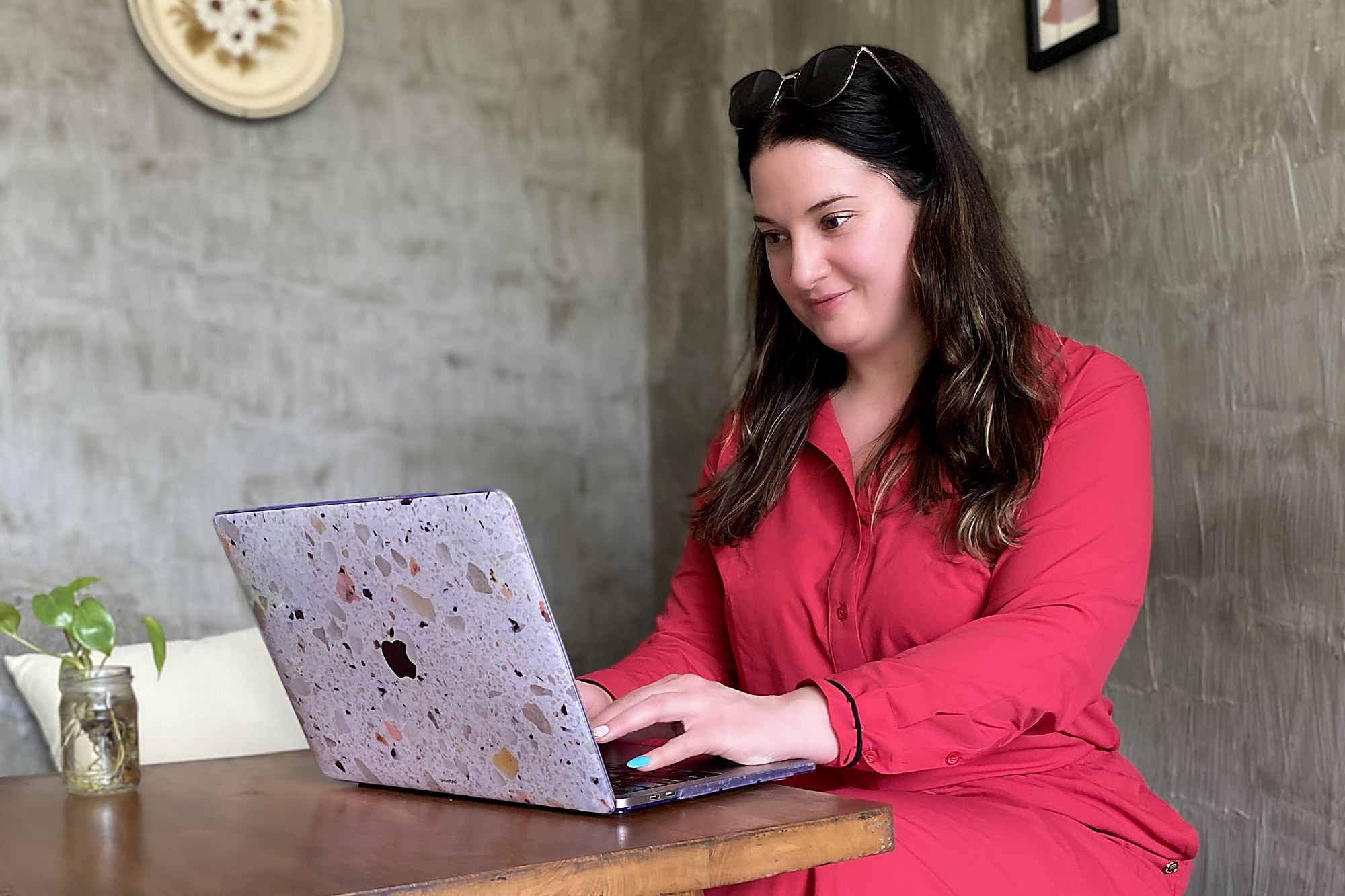 Marybeth Welty in a red dress working on her terrazzo-cased MacBook at a wooden table in an open-air Bali cafe
