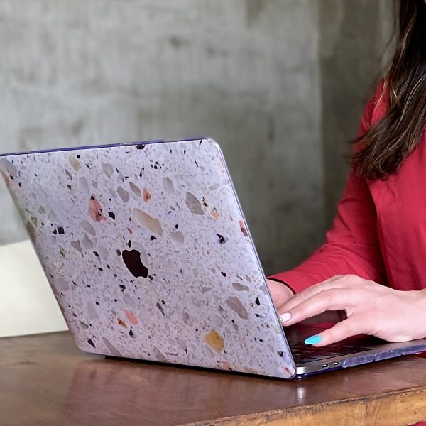 A terrazzo-cased laptop open on a wooden table beside a plant in a glass jar, hands typing