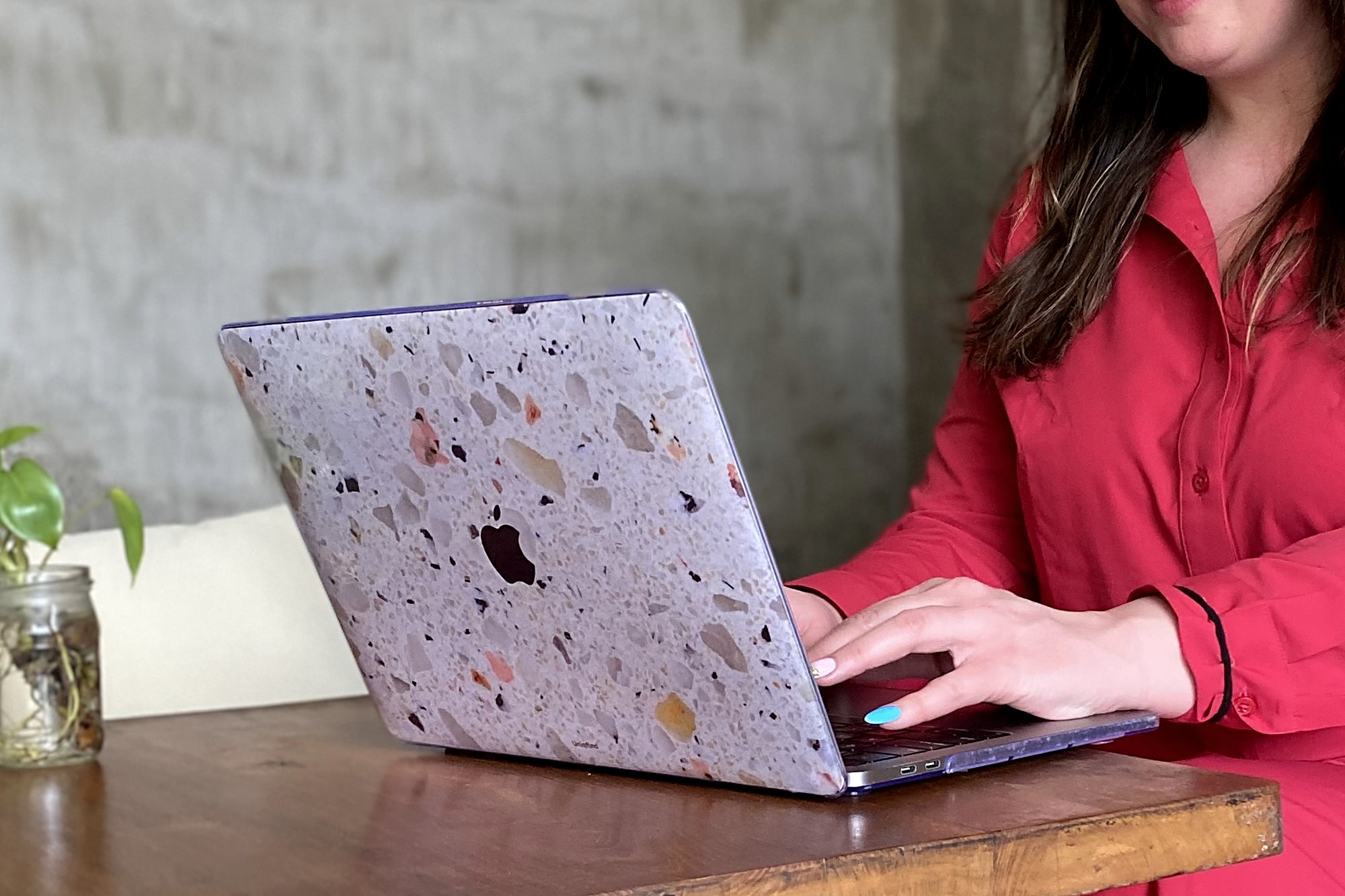 A terrazzo-cased laptop open on a wooden table beside a plant in a glass jar, hands typing